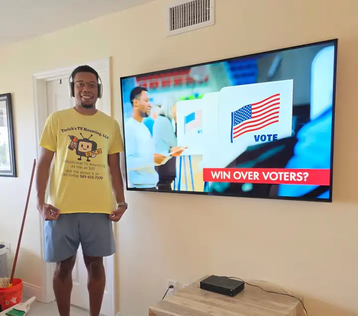 Tarick, owner of Tarick's TV Mounting, next to a Samsung TV mounted on a living-room wall in Melbourne, FL, with the power cord hidden inside the wall