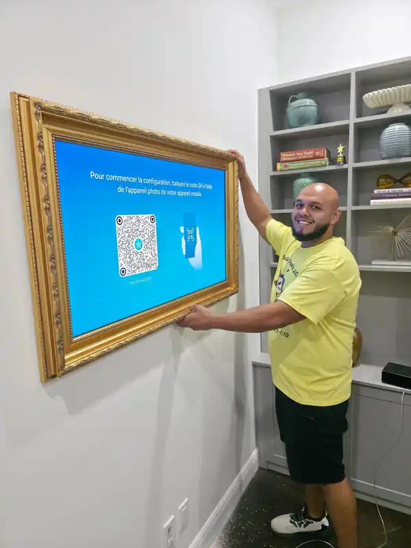 Noel, a Tarick's TV Mounting technician, holding a Samsung The Frame TV in a gold frame mounted on a wall in Palm Beach Gardens, FL